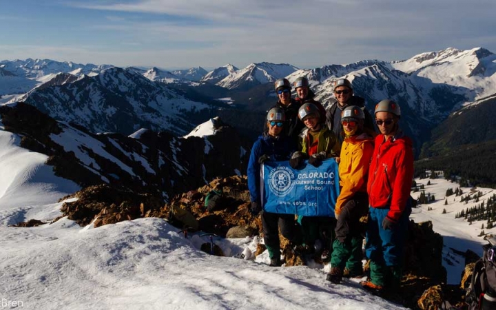 A group of gap year students wearing helmets hold an Outward Bound flag as they stand atop a snowy summit. There are vast snowy mountains in the background.
